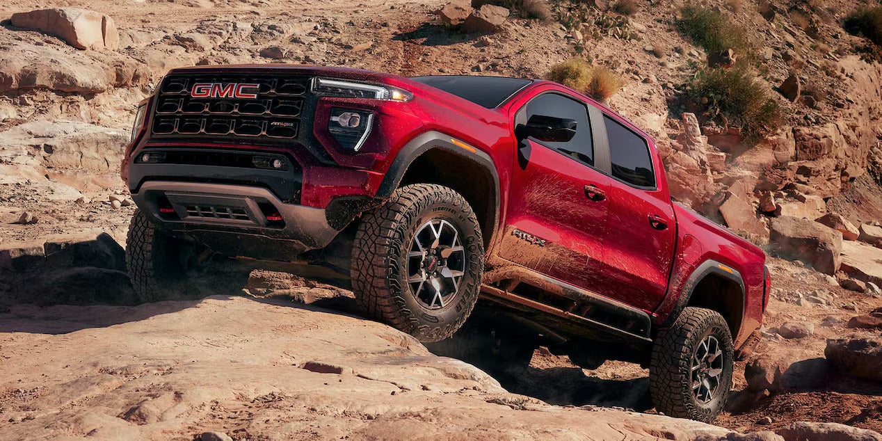 a red gmc canyon driving over large rocks in the desert