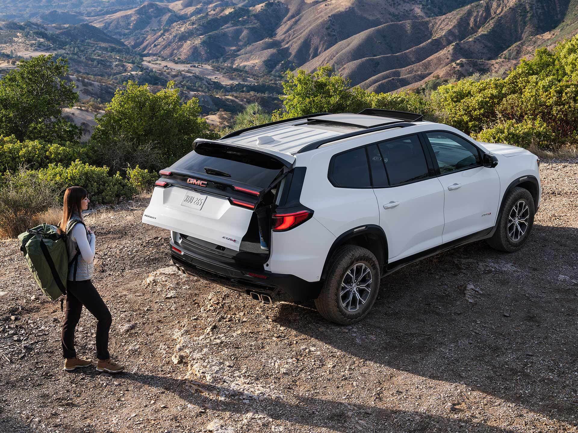 a white gmc acadia parked overlooking a cliff with the back open slightly and a woman walking towards it.