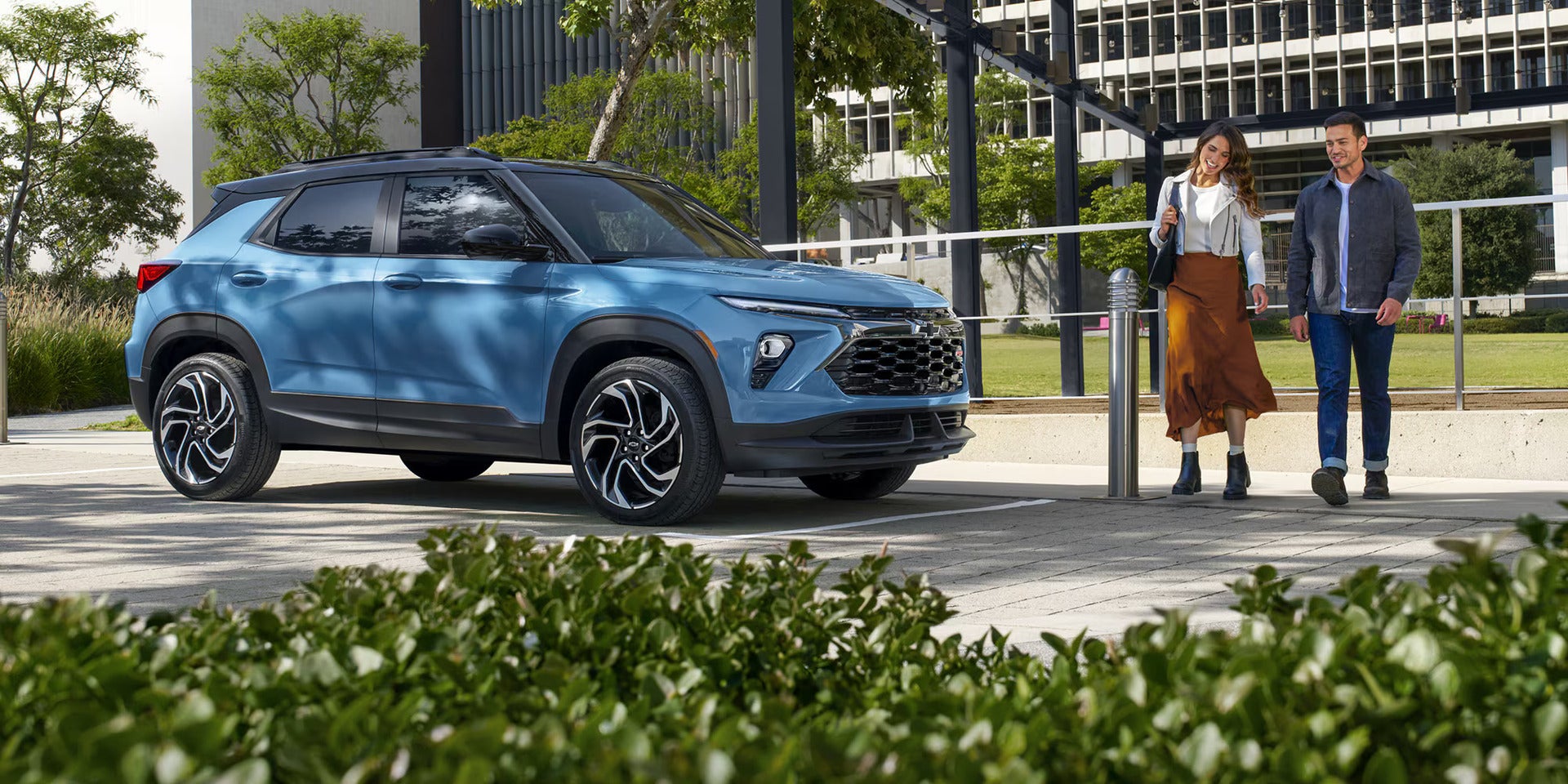 a blue trailblazer parked next to an office building with 2 people walking towards the vehicle.